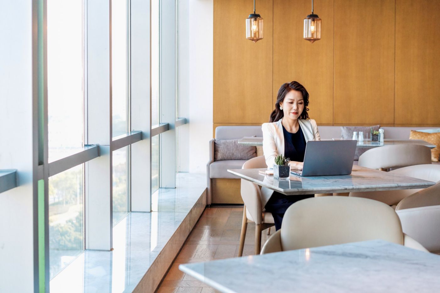 A woman in professional attire works on her laptop at a sunlit marble table in a modern corporate lounge.