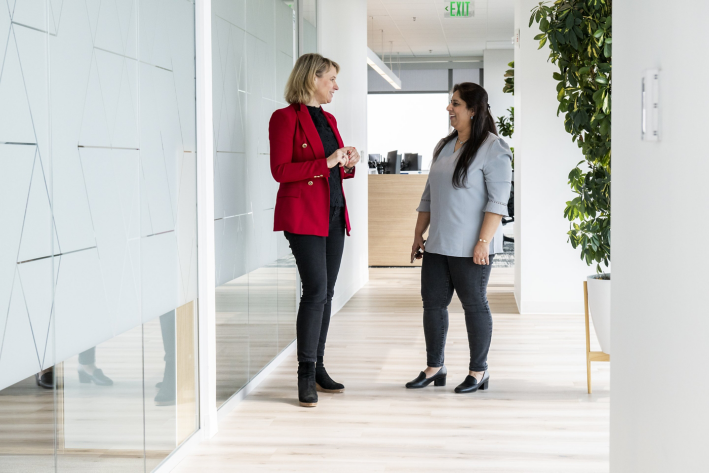 Two female colleagues engage in a friendly, professional conversation within a bright, modern corporate office hallway.