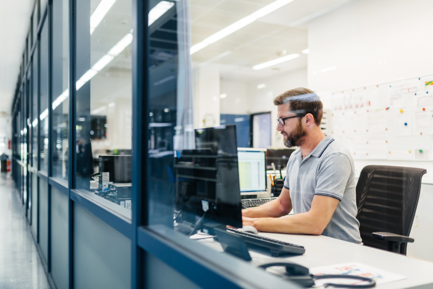 A focused male employee in a modern office uses a computer while seen through a glass partition.