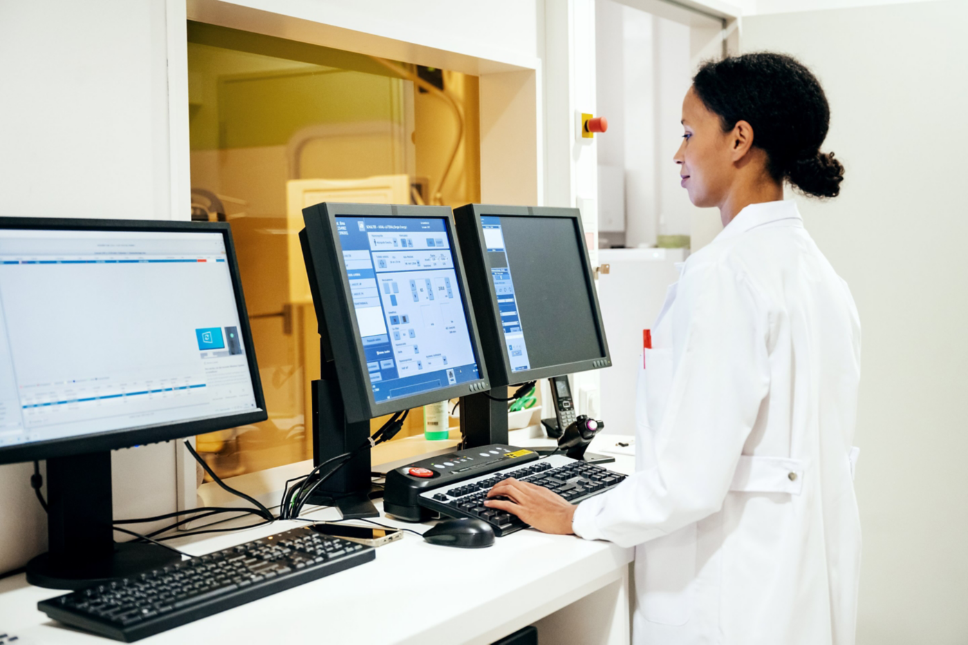 A professional radiologist in a white lab coat monitors medical imaging data at a computer workstation in a clinical diagnostic environment.
