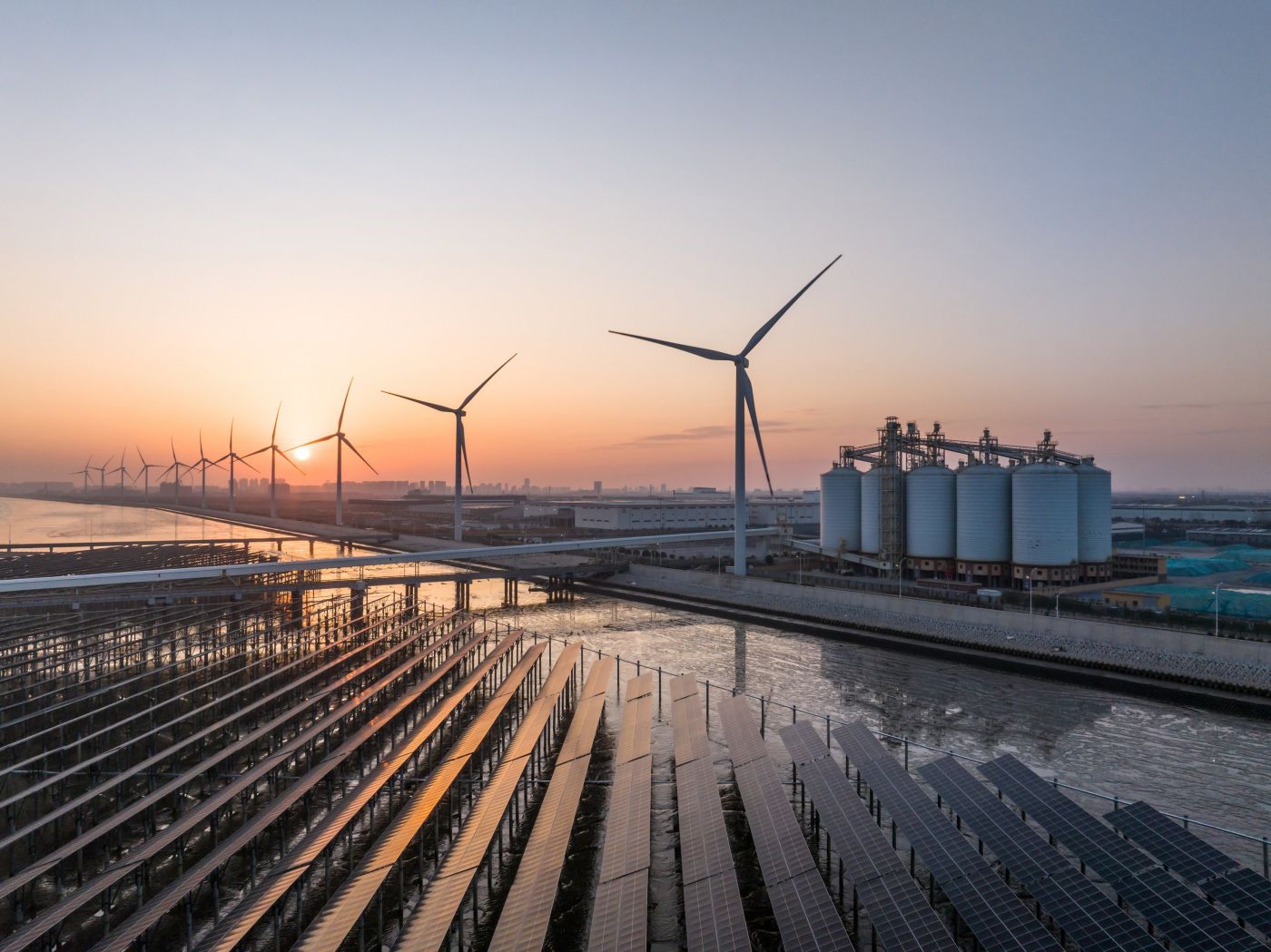 Solar panels and wind turbines capture energy alongside industrial storage silos at sunrise.