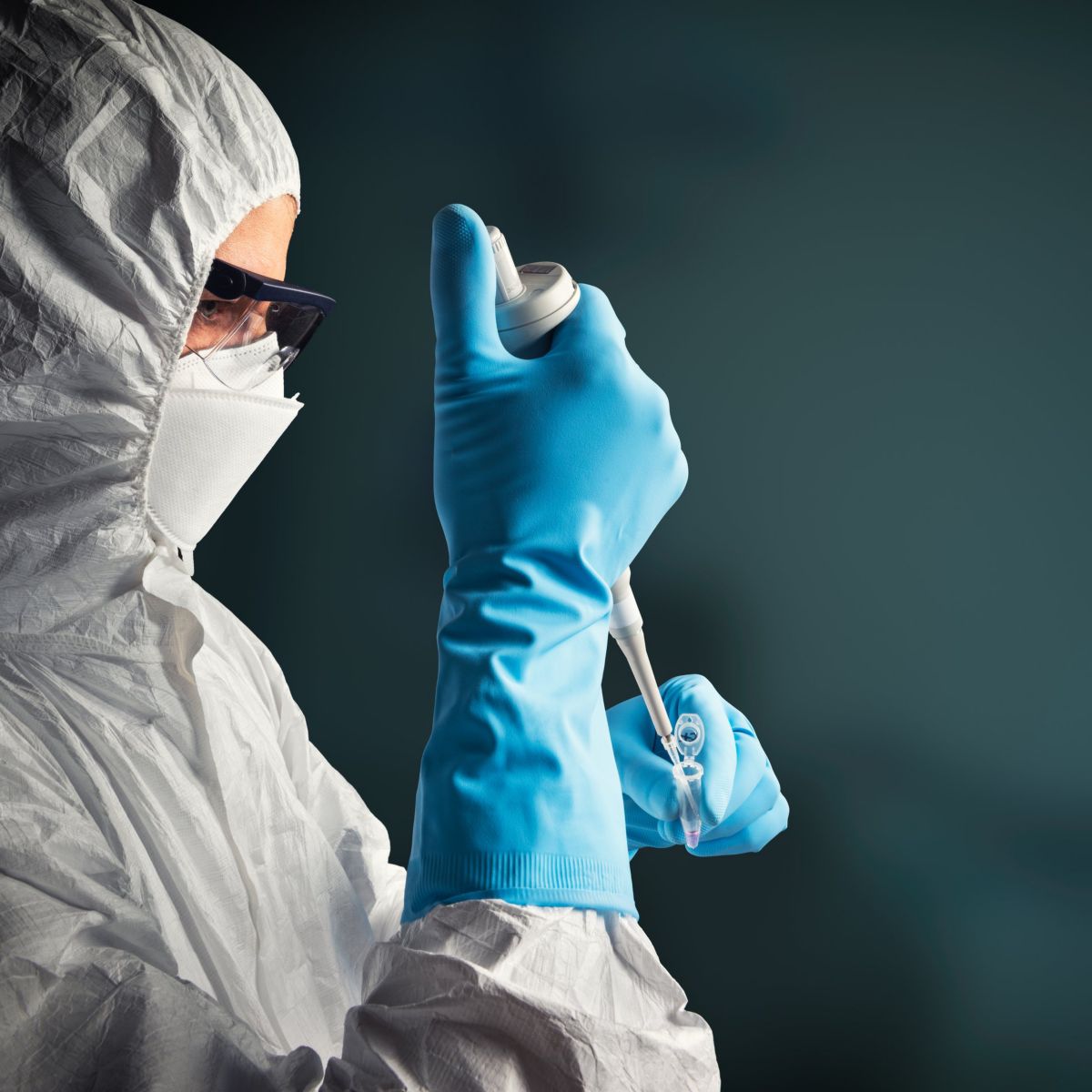 A researcher in protective gear uses a laboratory pipette to carefully extract a sample into a small vial against a dark background.