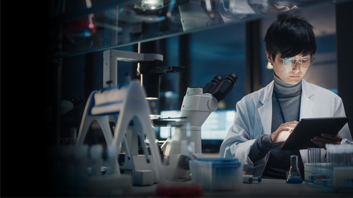A scientist works with a tablet beside a microscope and laboratory equipment in a dark research space.