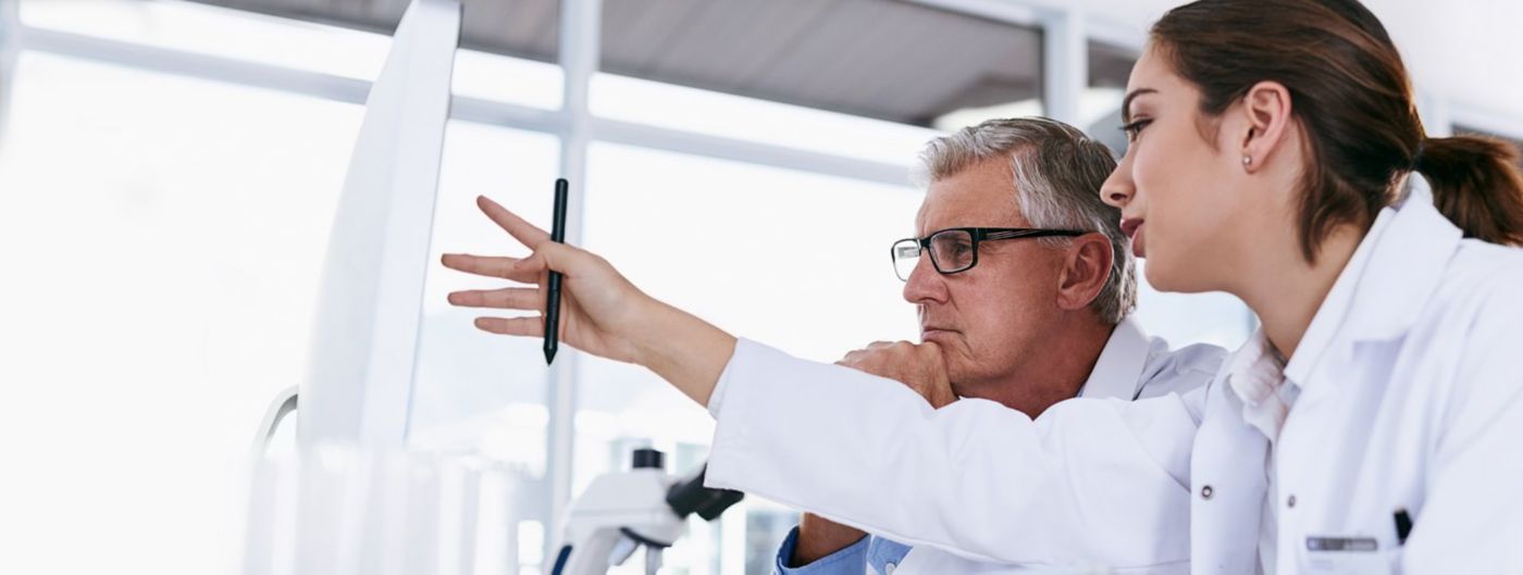 Two laboratory scientists discuss findings while one gestures toward a bright display or whiteboard.