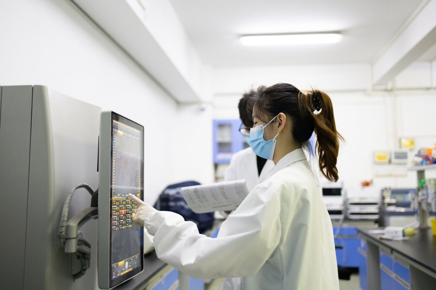 Two researchers in lab coats and masks operate an analytical instrument in a clean, professional research facility.