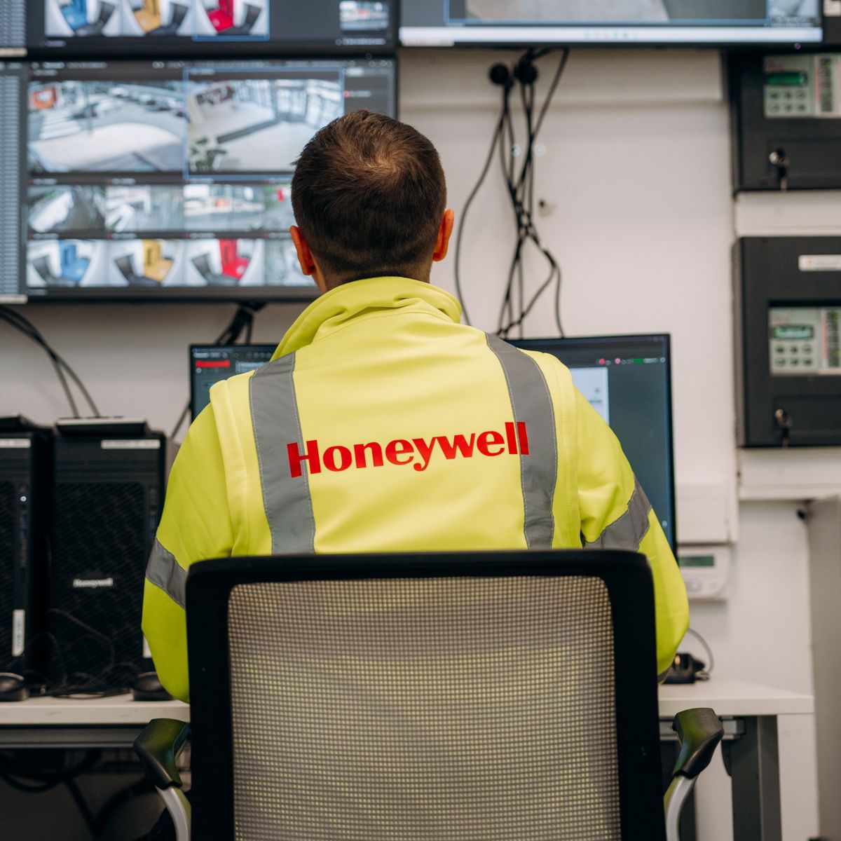A technician in a high-visibility jacket monitors multiple surveillance screens within a corporate security control room.