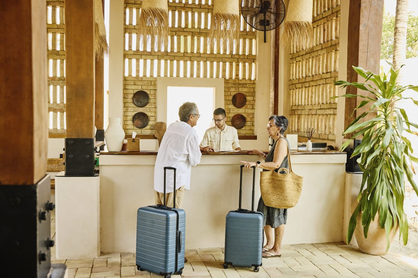 A senior couple stands at a resort reception desk with luggage, interacting with a staff member in a warm, naturally lit tropical lobby.