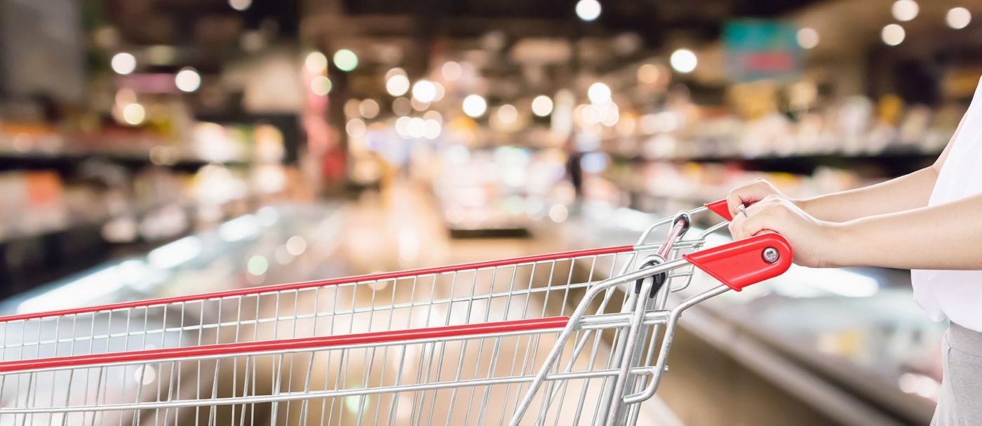 A red-handled shopping cart stands in a brightly lit supermarket with shelves and displays blurred in the background.