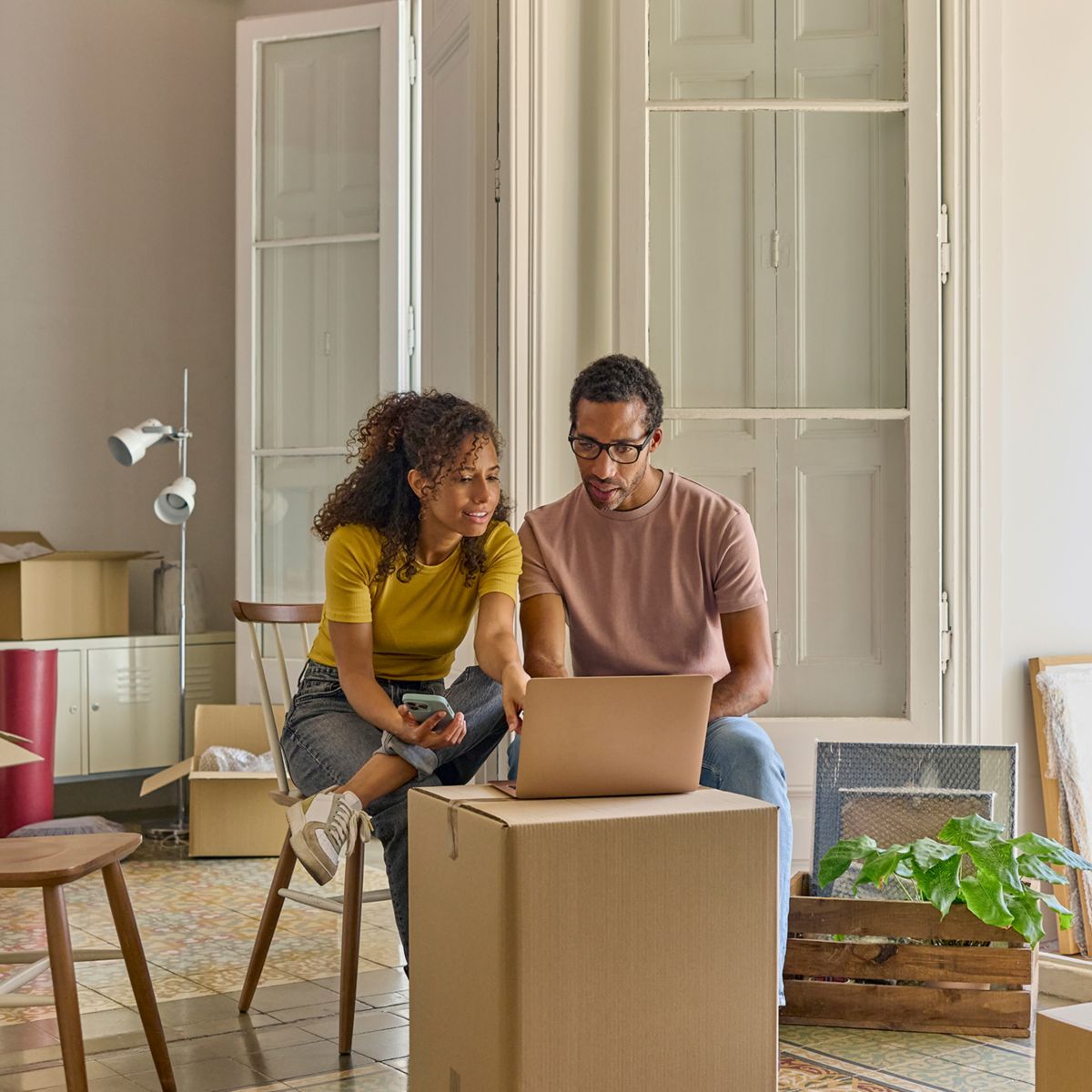 Two people sitting with a laptop and phone among moving boxes in a new home