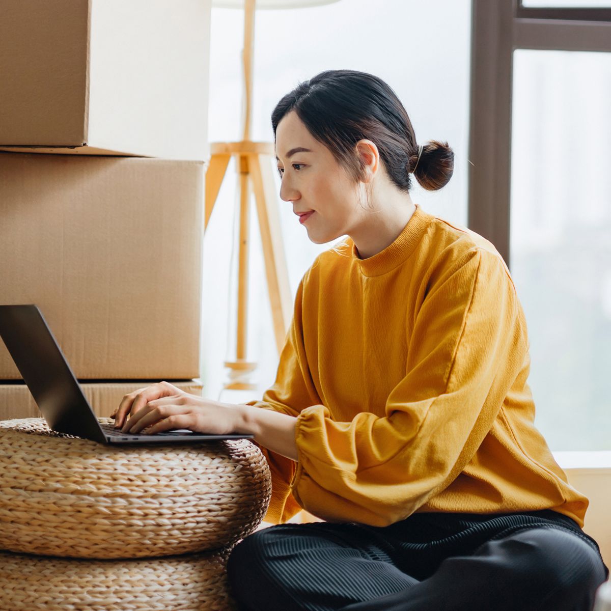 Person using a laptop while sitting on a stool surrounded by moving boxes in a home