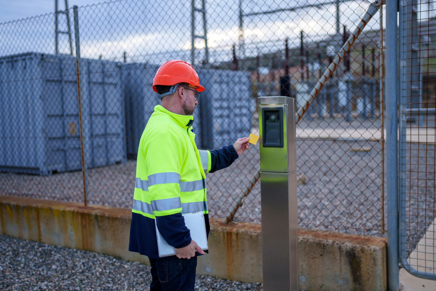 High-visibility jacket–clad utility worker holding a laptop swipes a key card at an access control reader beside a chain-link fence at an industrial site.