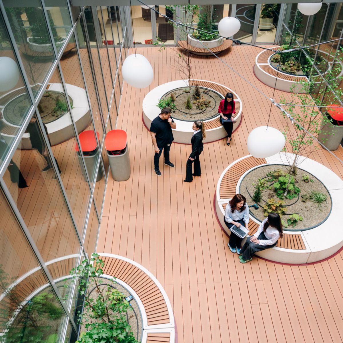 Overhead view of an indoor office atrium with people standing and sitting, circular planters, wooden flooring, and glass walls.