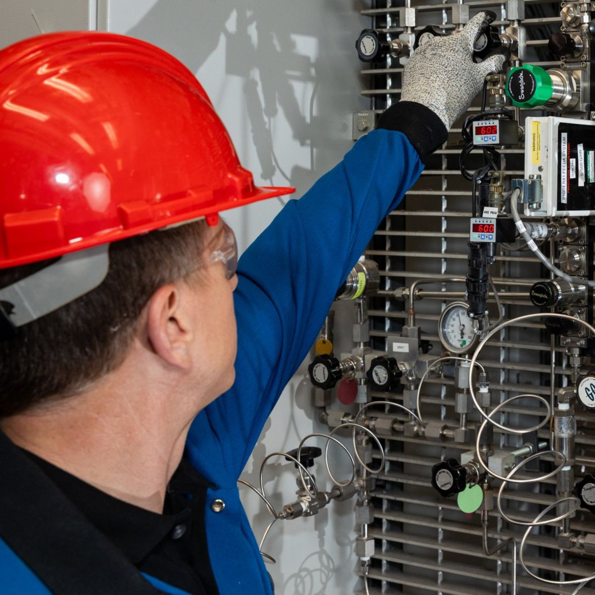 A technician in protective gear manually adjusts a valve on a complex industrial control panel featuring gauges, sensors, and intricate piping.