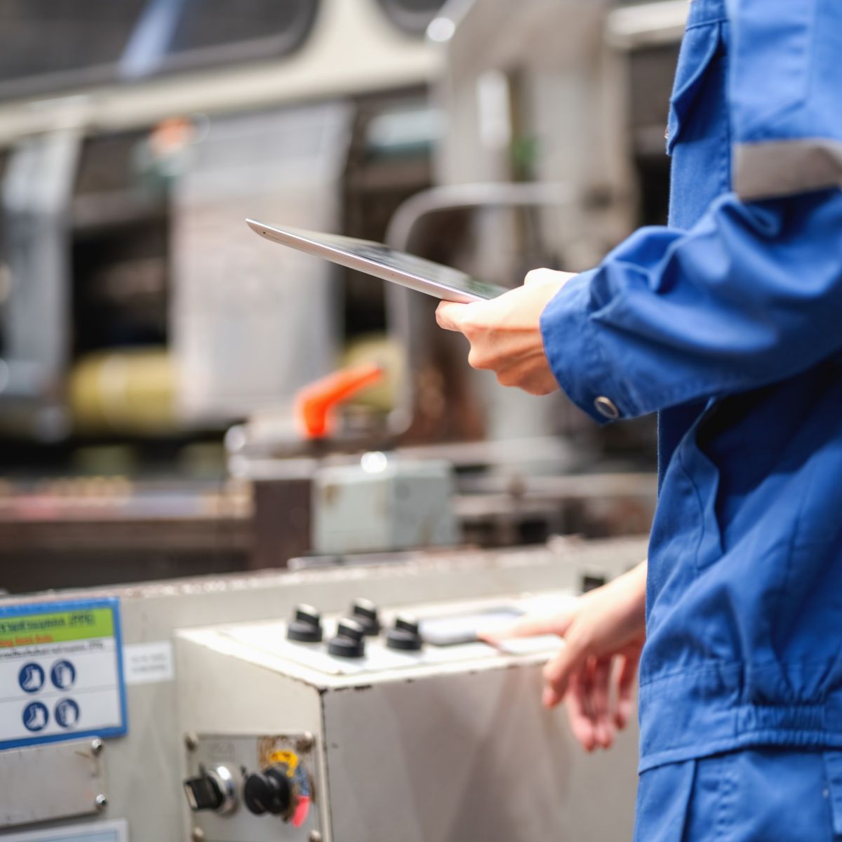 A technician in blue workwear uses a tablet to monitor and control industrial printing equipment in a factory setting.
