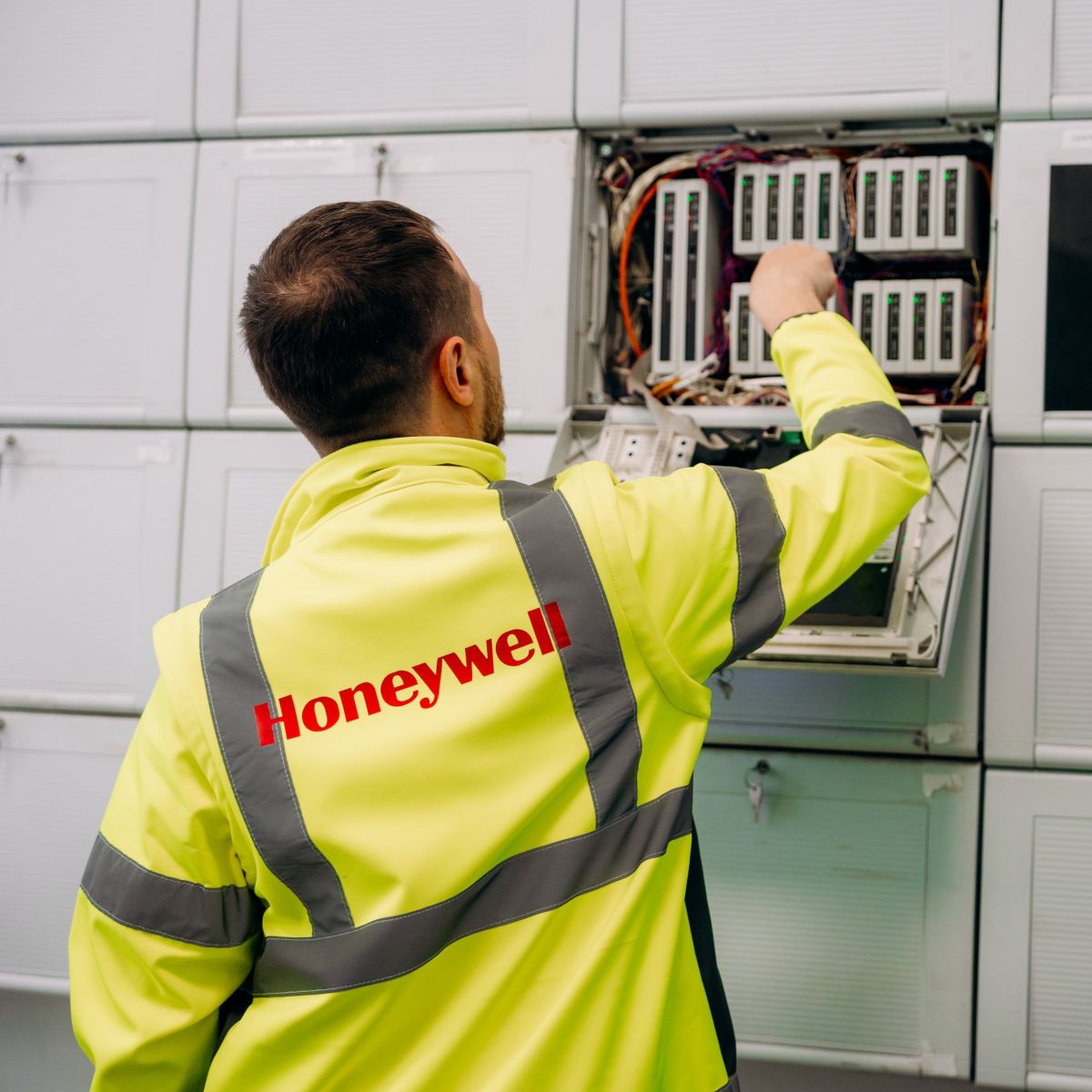A Honeywell technician wearing a high-visibility jacket performs maintenance on an industrial electronic control panel in a technical server room.