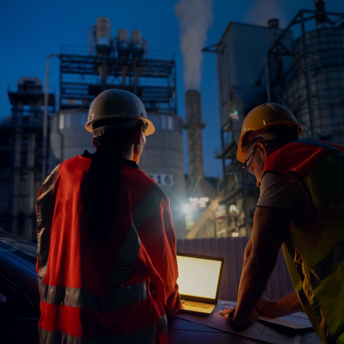 Engineers monitoring an industrial facility at night using laptop and field data, ensuring system performance, safety, and efficiency.
