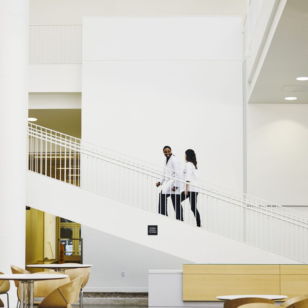 Hospital interior space with white walls, stairway railings, tables, chairs, and two people walking along a staircase.