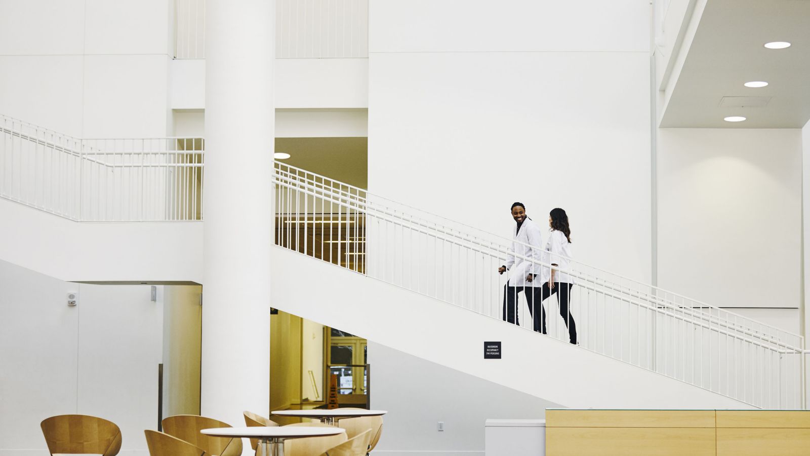 Hospital interior space with white walls, stairway railings, tables, chairs, and two people walking along a staircase.