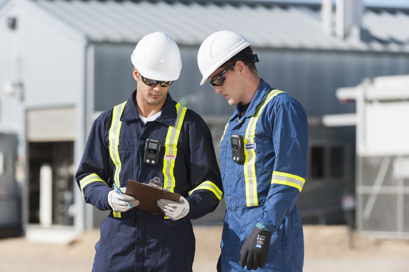 Two workers wearing helmets, a BW Clip 5 gas monitor and protective clothing reviewing notes on a clipboard, standing outdoors at an industrial site with equipment in the background.
