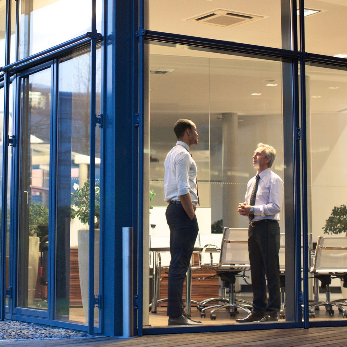 Two office colleagues meeting outside a modern glass office building, highlighting professional collaboration in a corporate environment.
