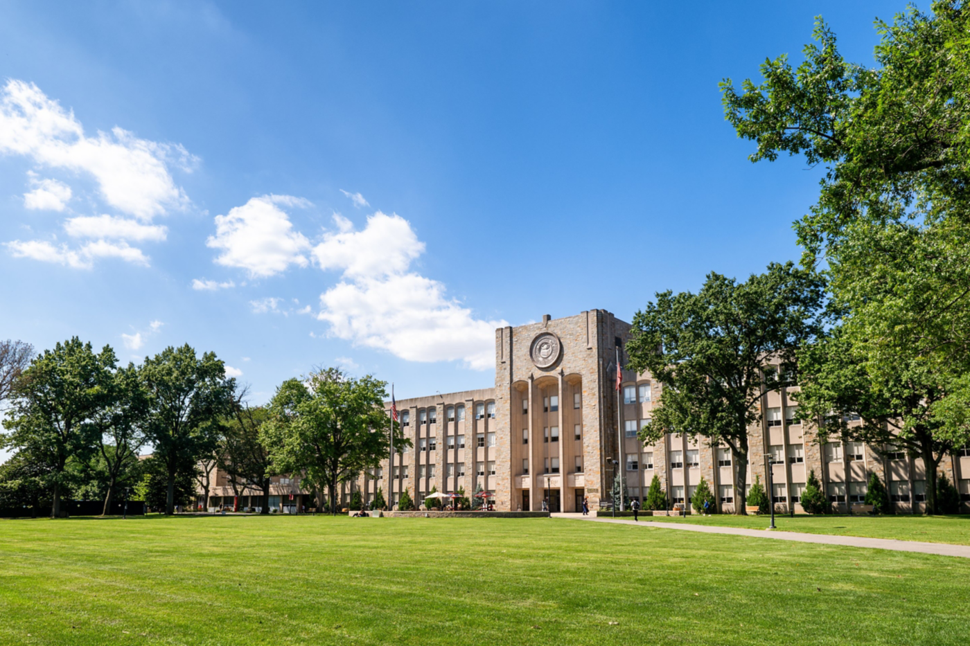 A stone collegiate building stands prominently behind a manicured green lawn under a bright blue sky.