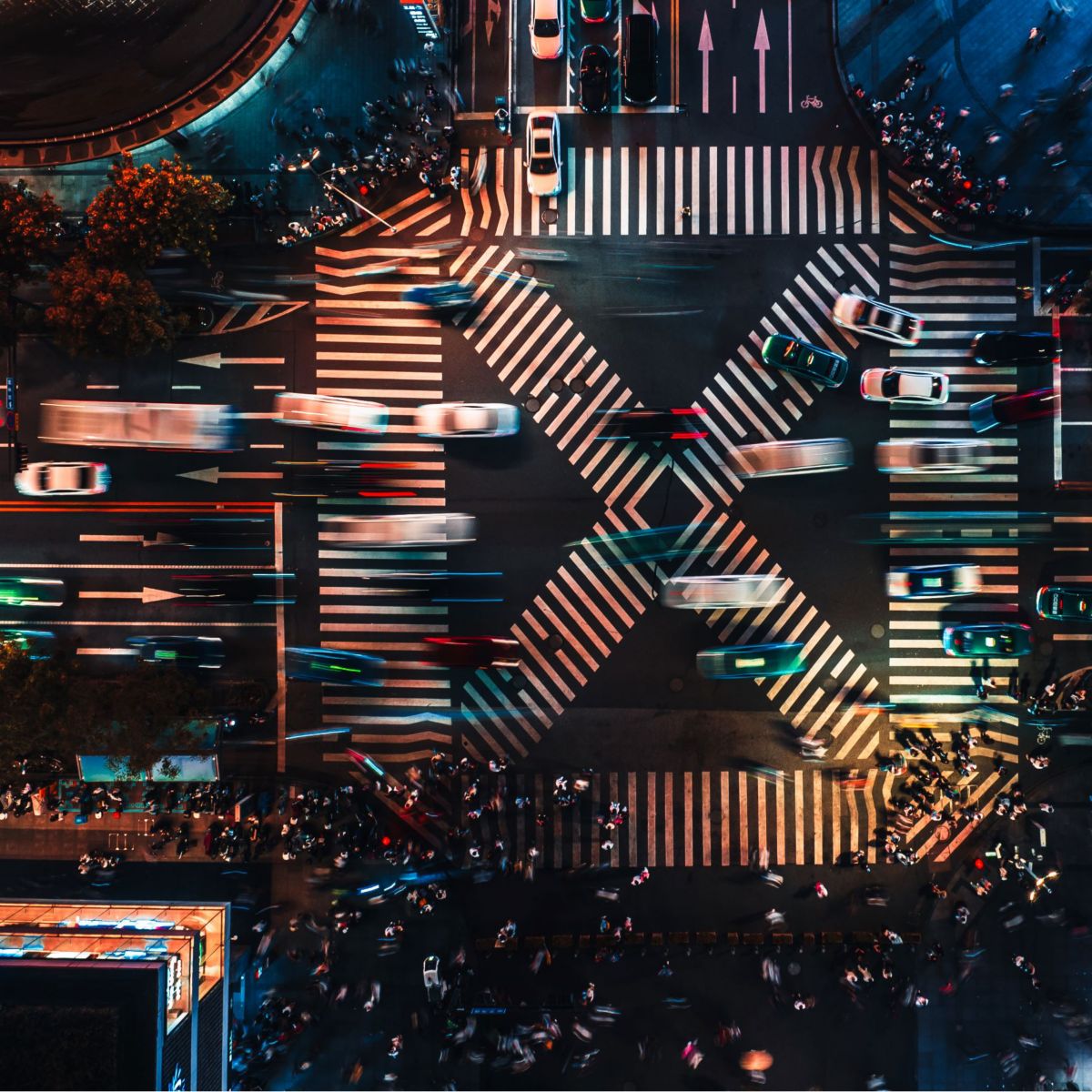 A bustling urban intersection at night showcases a mix of moving vehicles and pedestrians in vibrant city lights.