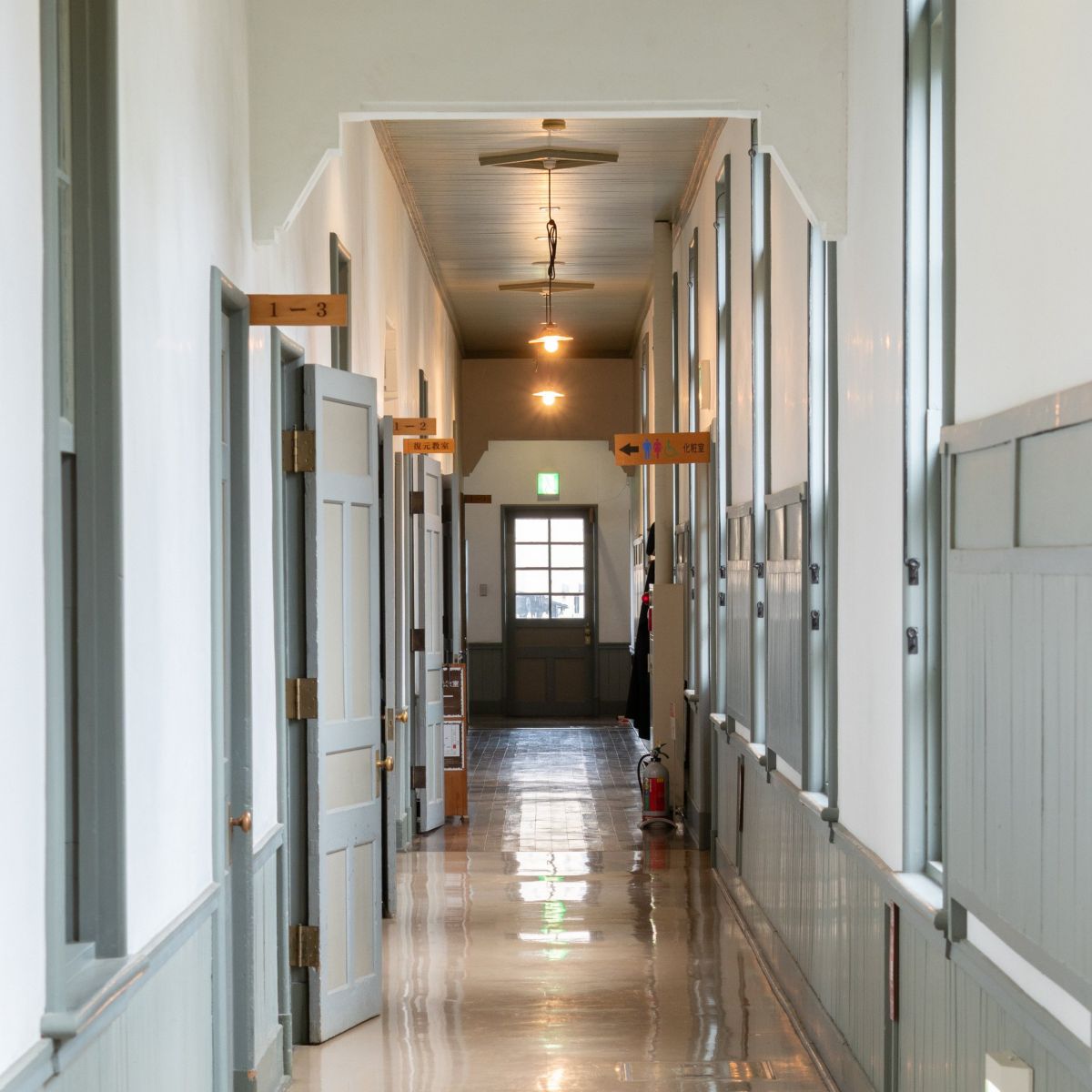 A symmetrical perspective shot down a narrow, sunlit hallway in a vintage wooden school building with reflective polished floors and open classroom doors.