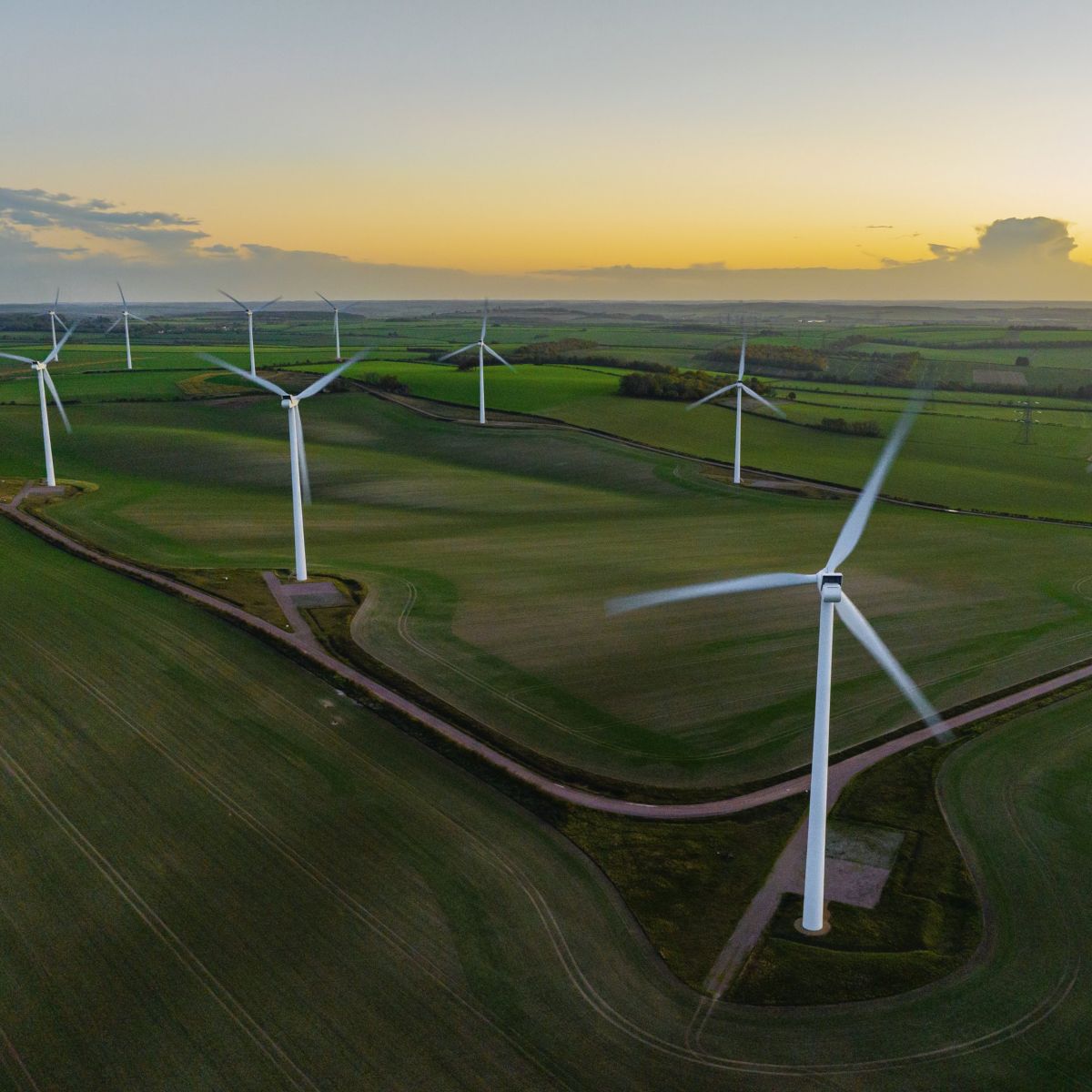 An aerial view of wind turbines in a lush green countryside at sunset, highlighting renewable energy infrastructure against a fading evening sky.