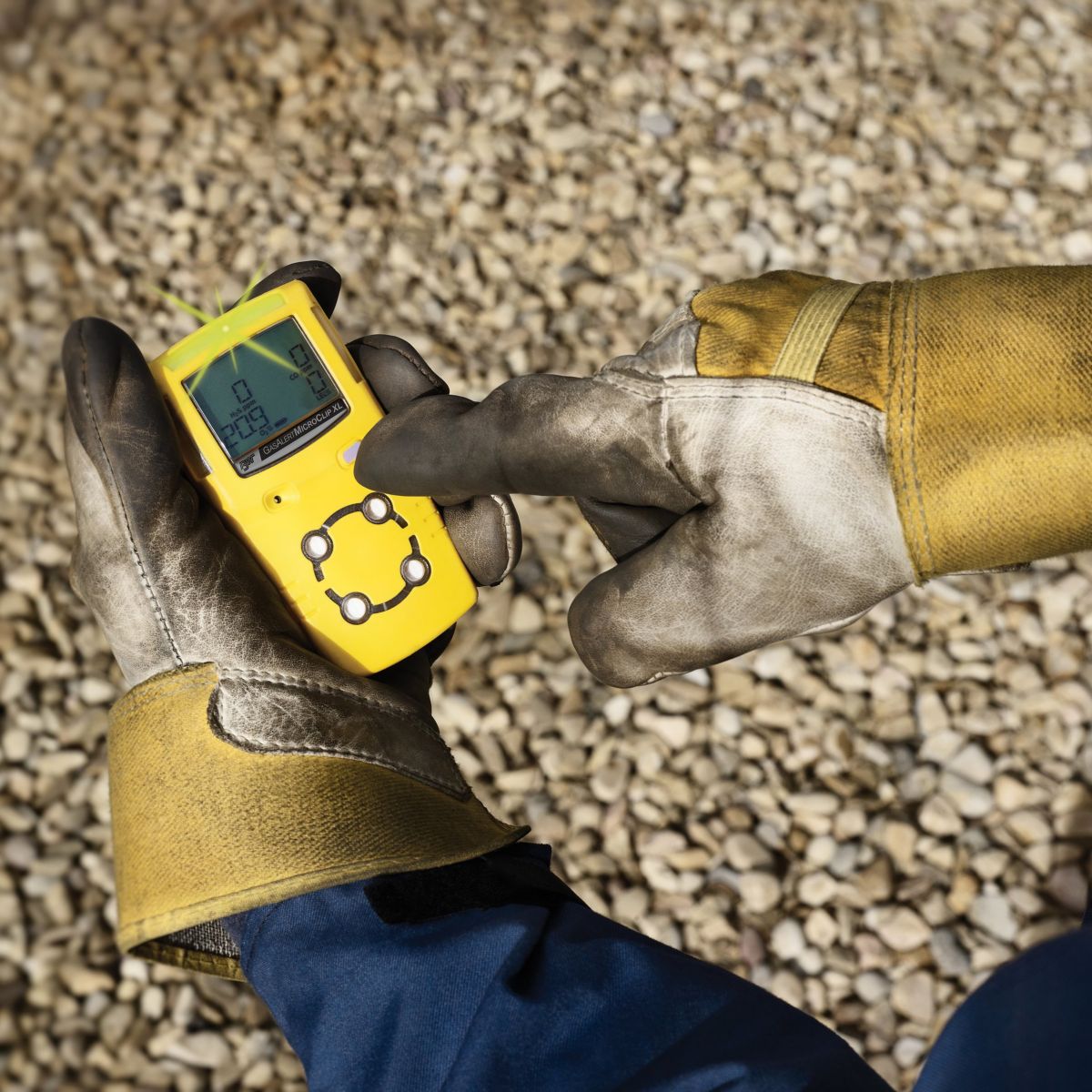 A worker wearing heavy-duty safety gloves monitors a handheld gas detector device over a gravel worksite.