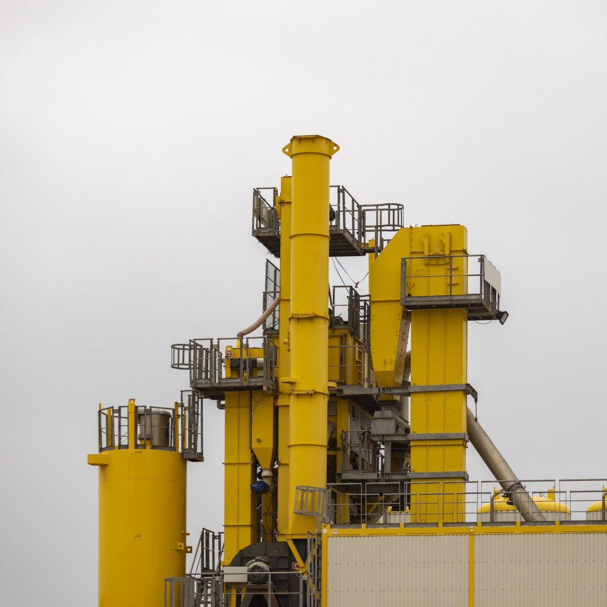 A bright yellow industrial processing facility stands against an overcast sky, highlighting its steel structure, vertical silos, and maintenance platforms.