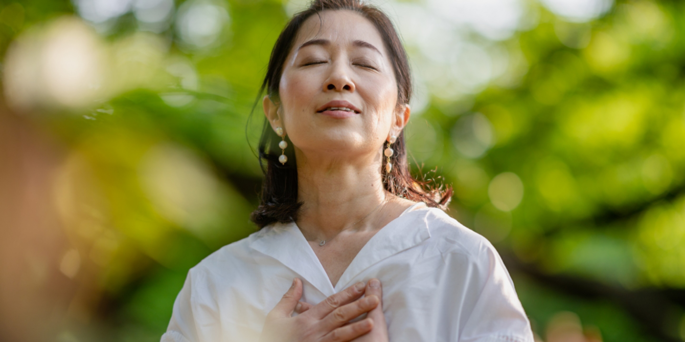 A woman is closing her eyes, doing breathing exercise and meditating in nature.