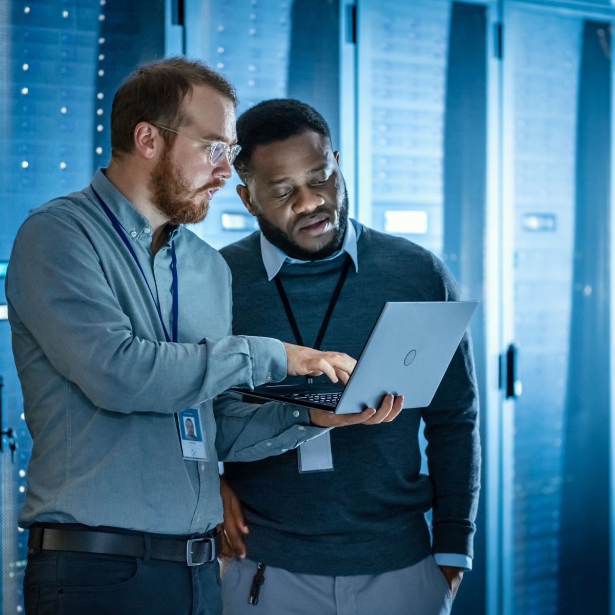 Bearded IT Technician in Glasses with Laptop Computer and Black Male Engineer Colleague are Using Laptop in Data Center while Working Next to Server Racks. Running Diagnostics or Doing Maintenance Work