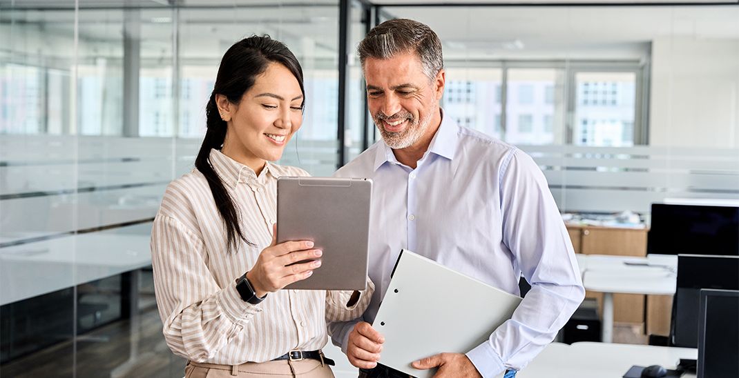 Two happy professional business people team Asian woman and Latin man workers working using digital tablet tech discussing financial market data standing at corporate office meeting.
